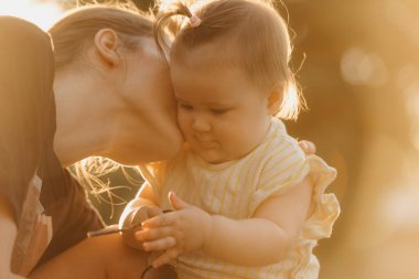 Sarı elbiseli bir bayan bebek güneş ışınlarında annesinin yanında güneş gözlükleriyle oynuyor. Anne gün batımında parkta küçük kızını öpüyor..