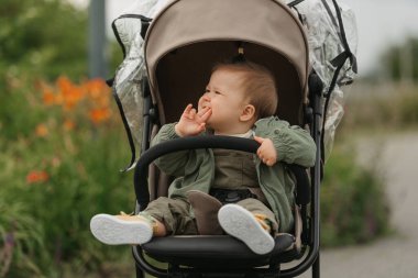 Dişi bir bebek, bulutlu bir günde parmaklarını ağzına sokup bebek arabasının yan tarafına bakıyor. Yeşil bir köyde, yağmurluklu güzel bir kız bebek arabasında..