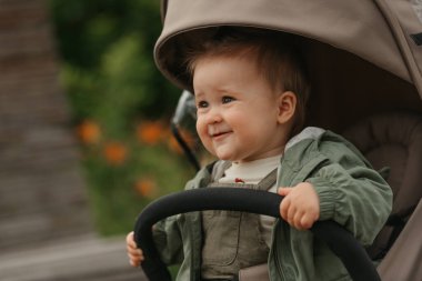 A close portrait of a female toddler who is holding the safety bumper bar of the stroller on a cloudy day. A young girl in a raincoat is in her baby carriage in a park at noon.