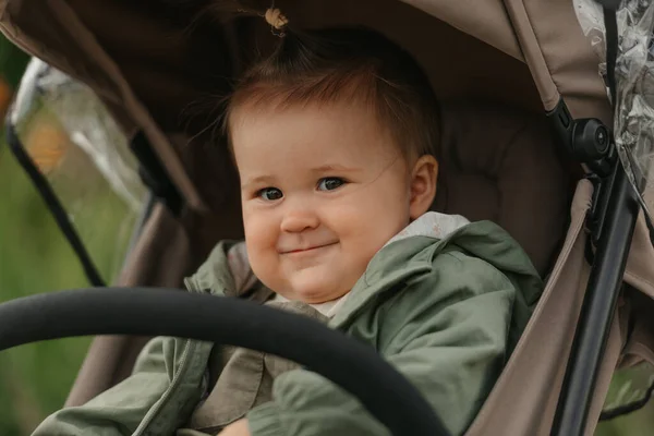 A close portrait from the side of a female toddler who is posing in the stroller on a cloudy day. A young girl in a raincoat with open eyes is in her baby carriage in a park at noon.