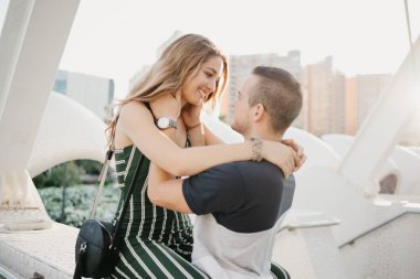 A close photo of a smiling girl who is hugging her boyfriend in the modern urban space at sunset in Spain. A couple of tourists on a date in the evening in Valencia.