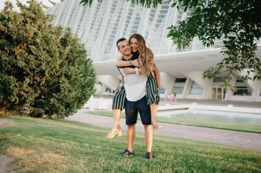 A full-length photo of a smiling couple of tourists who are having fun in the modern urban space on a date at sunset in Valencia. A girl is riding her boyfriend in the evening in Spain.