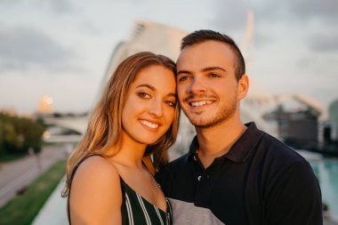 Bir çift gülümseyen turist, arka plandaki modern kentsel alanla Valencia 'da gün batımında eğleniyor. Bir adam akşam İspanya 'da kız arkadaşına sarılıyor.