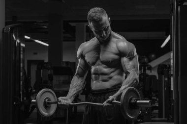 Sakallı bir vücut geliştirici spor salonunda EZ halteriyle biseps yapıyor. Spor yaparken çıplak kaslı bir adamın gövdesinin fotoğrafı..