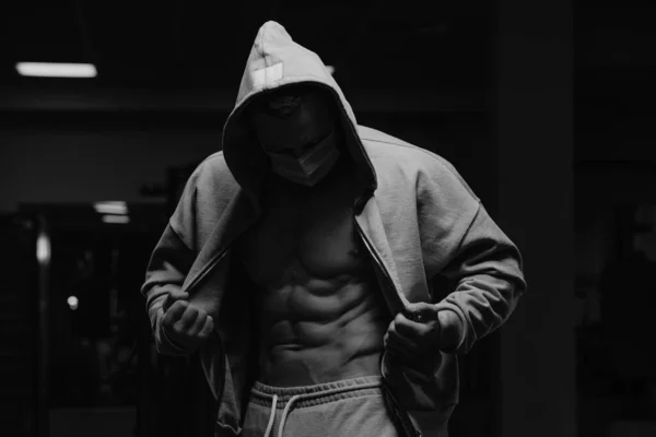 A bodybuilder in a hood and a face mask to avoid the spread of coronavirus is opening his zipped hoodie to demonstrate his abdominal muscles. A sporty guy in a surgical mask is posing after a workout
