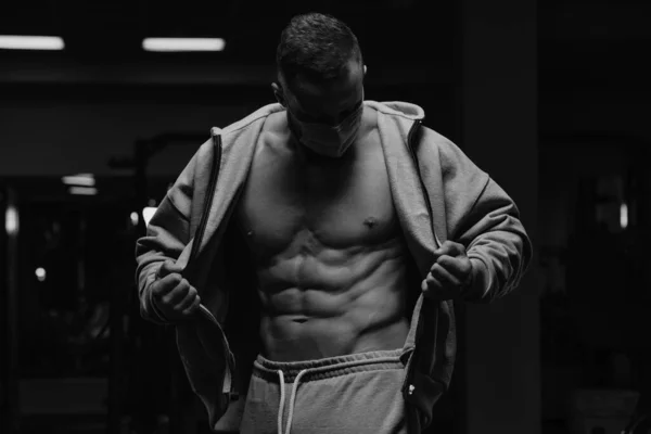 A bodybuilder in a face mask to avoid the spread of coronavirus is opening his zipped hoodie to demonstrate his abdominal muscles. A sporty guy in a surgical mask is posing after a workout in a gym.