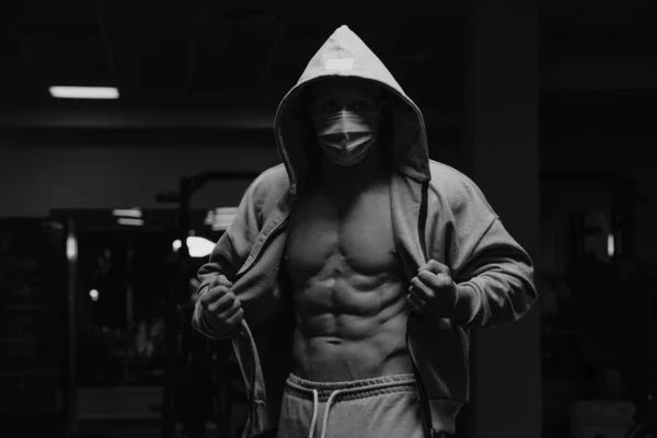 A bodybuilder in the hood and a face mask to avoid the spread of coronavirus is opening his zipped hoodie to demonstrate his abs. A sporty guy in a surgical mask is posing after a workout in a gym.