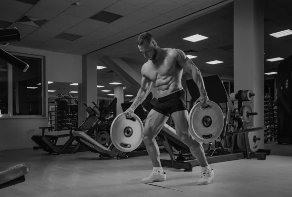 A bodybuilder in shorts is walking through the gym with two rubber plates. A shirtless sporty guy is training alone.