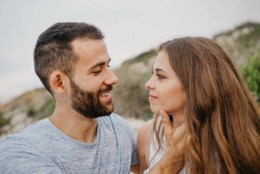 A close photo of a smiling Hispanic man who is stroking with his hands the face of his Latina girlfriend on the rocky sea coast in Spain. A couple of tourists in the evening on the hills in Valencia clipart