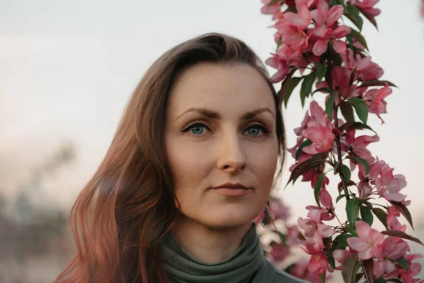 A headshot of a redhead woman is staring into the distance near flowering sakura at sunset.