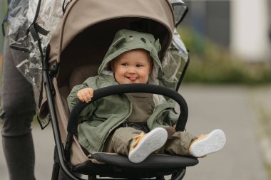 A female toddler with her tongue out is sitting in the stroller on a cloudy day. A young girl in a baby carriage in a village green.
