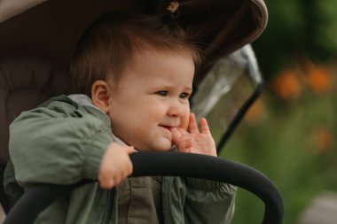 Bulutlu bir günde parmaklarını bebek arabasının ağzına sokan bir kız çocuğunun yakın portresi. Yeşil bir köyde, yağmurluklu genç bir kız bebek arabasında..