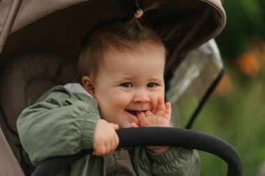 Bebek arabasında parmaklarını ağzına sokan küçük bir kızın yakın portresi. Yağmurluk giymiş genç bir kız öğle vakti bir parkta bebek arabasında..