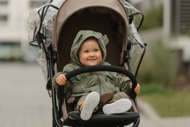 A female toddler is holding the safety bumper bar of the stroller on a cloudy day. A young girl in the hood of her raincoat is in her baby carriage in a park at noon.
