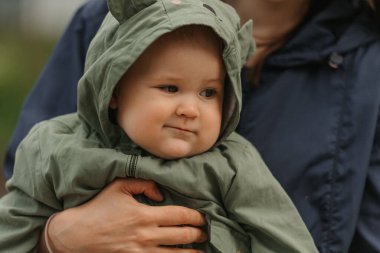 A close side photo of a female toddler who is carried by her mom in the street at noon. A mother is holding her young pretty daughter near their home on a cloudy day.