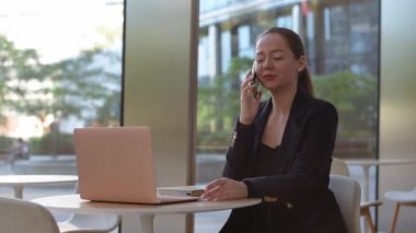 A female realtor in a blazer is canceling a conversation on the cell phone. A woman is sitting at the table in the modern lobby. A female IT developer near a laptop in a contemporary office interior.