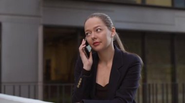 A female realtor is calling to her colleague while leaning on the parapet in a modern urban space in the financial district. A businesswoman in the contemporary garden in a cluster of tall buildings.