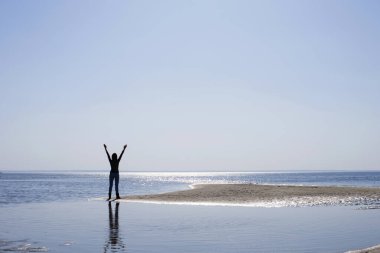 Nehir kenarında güneşin altında kolları açık bir kadın silueti.