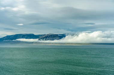 Antlantik okyanusun İzlanda kıyı şeridindeki renkli manzarası, turkuaz su, arka planda dağlar ve alçalan beyaz bulutlar..