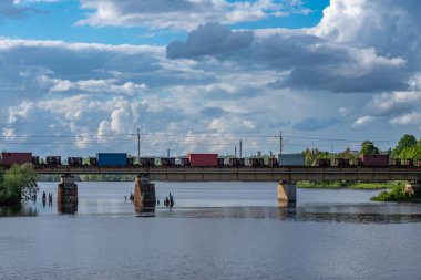 Kargo ya da yük treni yaz güneşinde beyaz kabarık bulutlarla köprüden geçiyor.