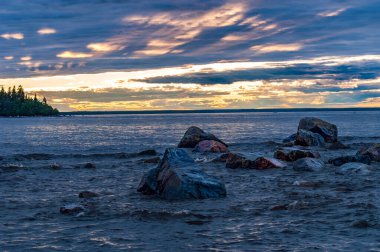 Deniz kıyısındaki suda kayalar Güneş bulutların arasından batarken