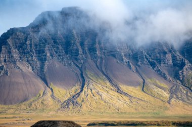 Taşlaşmış lav akıntılarıyla dolu büyük bir dağ zirvesi manzarası. Beyaz bulut kısmen tepeyi kaplıyor. Dağın eteğindeki küçük eve dikkat edin.