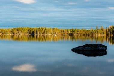 İsveç 'te küçük bir gölün karşısındaki yeşil bir ormanın güzel yaz manzarası. Gökyüzündeki bulutlar sakin suları yansıtırken