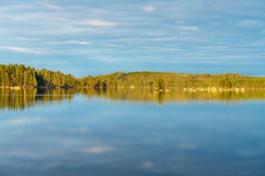 İsveç 'te bir gölün karşısındaki yeşil bir ormanın güzel yaz manzarası. Gökyüzündeki bulutlar sakin suları yansıtırken