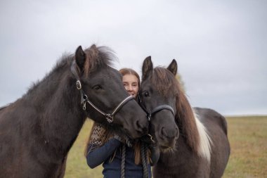 Genç bir kadın tarlada dikilip en iyi iki arkadaşına sarılıyor, İzlanda atlarına.