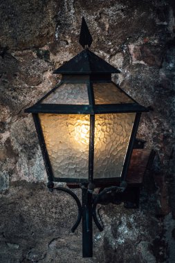 Taş bir duvara asılmış metal ve camdan yapılmış eski bir lamba.