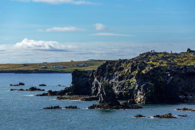 İzlanda 'nın batı kıyısından yaz manzarası, yaz güneşi ve mavi bir gökyüzü. Arka planda küçük bir köy ve lav oluşumunun üstünde yürüyen turistler.