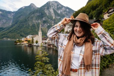 Hallstatt, Avusturya: Güzel romantik kadın turistin portresi, doğal ışık, vücut pozitifliği, bej renkli beyaz çizgili kız, şapka ve fular, manzara resimli şehir sokağı