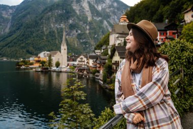 Hallstatt, Avusturya: Güzel romantik kadın turistin portresi, doğal ışık, vücut pozitifliği, bej renkli beyaz çizgili kız, şapka ve fular, manzara resimli şehir sokağı