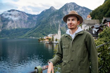 Hallstatt, Avusturya: Güzel romantik bir genç adamın portresi turist gülümsemesi, doğal ışık, bej şapkalı çocuk yeşil ceket, manzara manzaralı kasaba sokağı, göl kenarındaki dağ köyü, yaz günü Alpler