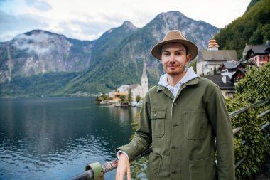 Hallstatt, Avusturya: Güzel romantik bir genç adamın portresi turist gülümsemesi, doğal ışık, bej şapkalı çocuk yeşil ceket, manzara manzaralı kasaba sokağı, göl kenarındaki dağ köyü, yaz günü Alpler
