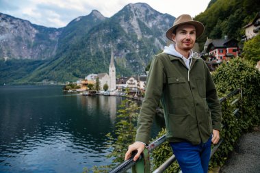 Hallstatt, Avusturya: Güzel romantik bir genç adamın portresi turist gülümsemesi, doğal ışık, bej şapkalı çocuk yeşil ceket, manzara manzaralı kasaba sokağı, göl kenarındaki dağ köyü, yaz günü Alpler