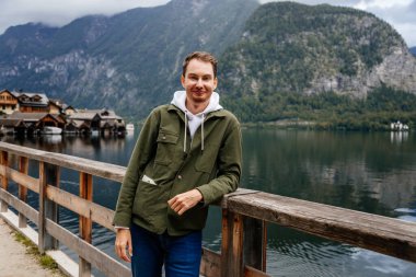 Hallstatt, Avusturya: Güzel romantik bir genç adamın portresi turist gülümsemesi, doğal ışık, bej şapkalı çocuk yeşil ceket, manzara manzaralı kasaba sokağı, göl kenarındaki dağ köyü, yaz günü Alpler