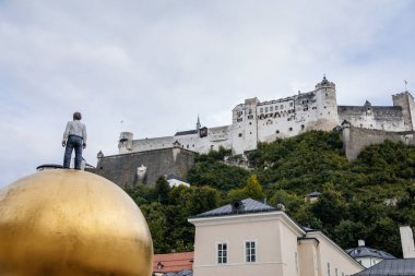Salzburg, Avusturya, 28 Ağustos 2021: Goldene Kugel heykelindeki adam, Sphaera heykeli, eski ortaçağ şatosu Festung Hohensalzburg, güneşli yaz gününde kuleleri olan güzel kale manzarası