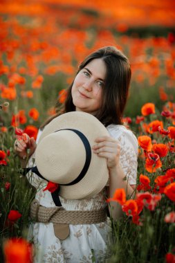 Beyaz ve bej elbiseli güzel bir kızın gün batımında haşhaş tarlasında saman şapkalı güzel bir kızın resmi, çiçekli romantik genç bir kadının portresi, pozitiflik, seçici doğal ışık.