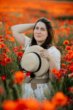 Beyaz ve bej elbiseli güzel bir kızın gün batımında haşhaş tarlasında saman şapkalı güzel bir kızın resmi, çiçekli romantik genç bir kadının portresi, pozitiflik, seçici doğal ışık.