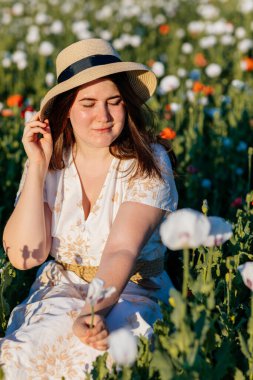 Beyaz ve bej elbiseli güzel bir kızın gün batımında haşhaş tarlasında saman şapkalı güzel bir kızın resmi, çiçekli romantik genç bir kadının portresi, pozitiflik, seçici doğal ışık.