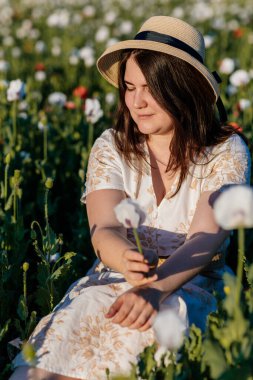 Beyaz ve bej elbiseli güzel bir kızın gün batımında haşhaş tarlasında saman şapkalı güzel bir kızın resmi, çiçekli romantik genç bir kadının portresi, pozitiflik, seçici doğal ışık.