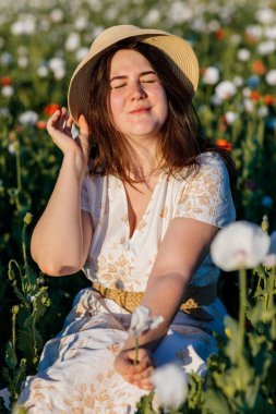 Beyaz ve bej elbiseli güzel bir kızın gün batımında haşhaş tarlasında saman şapkalı güzel bir kızın resmi, çiçekli romantik genç bir kadının portresi, pozitiflik, seçici doğal ışık.