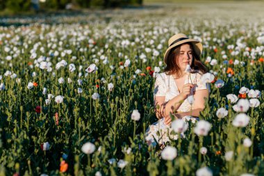 Beyaz ve bej elbiseli güzel bir kızın gün batımında haşhaş tarlasında saman şapkalı güzel bir kızın resmi, çiçekli romantik genç bir kadının portresi, pozitiflik, seçici doğal ışık.