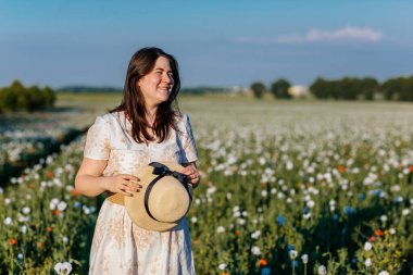 Beyaz ve bej elbiseli güzel bir kızın gün batımında haşhaş tarlasında saman şapkalı güzel bir kızın resmi, çiçekli romantik genç bir kadının portresi, pozitiflik, seçici doğal ışık.