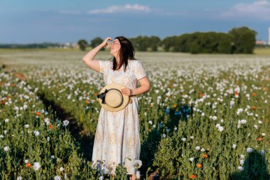 Beyaz ve bej elbiseli güzel bir kızın gün batımında haşhaş tarlasında saman şapkalı güzel bir kızın resmi, çiçekli romantik genç bir kadının portresi, pozitiflik, seçici doğal ışık.