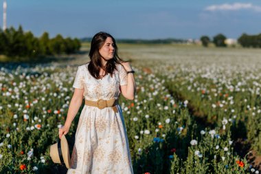 Beyaz ve bej elbiseli güzel bir kızın gün batımında haşhaş tarlasında saman şapkalı güzel bir kızın resmi, çiçekli romantik genç bir kadının portresi, pozitiflik, seçici doğal ışık.