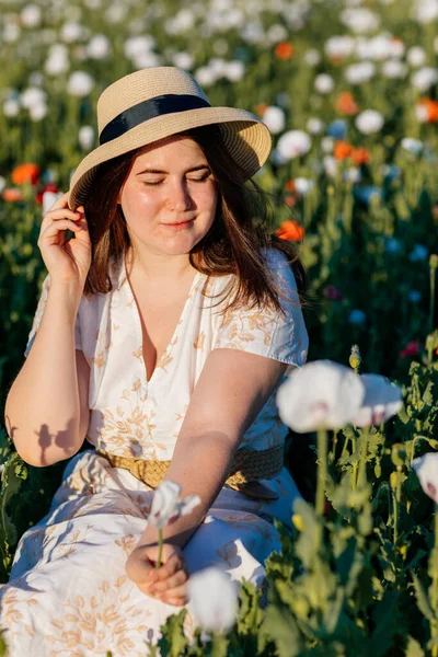 Beyaz ve bej elbiseli güzel bir kızın gün batımında haşhaş tarlasında saman şapkalı güzel bir kızın resmi, çiçekli romantik genç bir kadının portresi, pozitiflik, seçici doğal ışık.