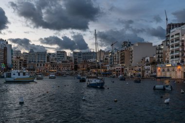 Saint Julian, Malta - 01 07 2022: Yat limanına, tatil dairelerine ve gün batımında teknelere ve renkli bulutlara bakın