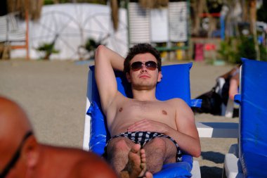 25-30 yaşlarında yakışıklı bir adam, esmer, ot gözlüklü, kumlu bir sahilde uzanmış, güneşleniyor, güneşleniyor.